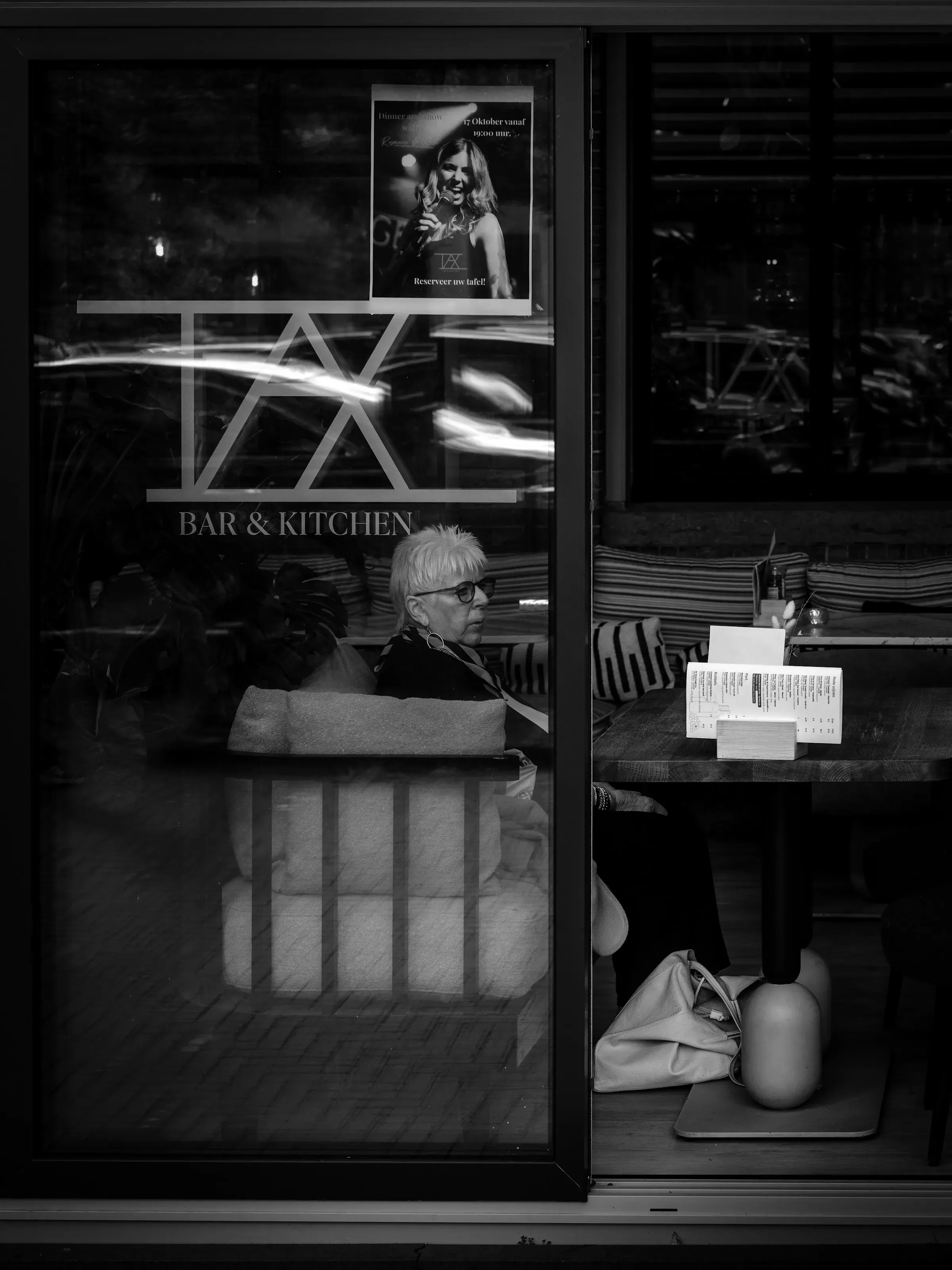 A woman seen through a café window, the glass carrying the reflection of the street.