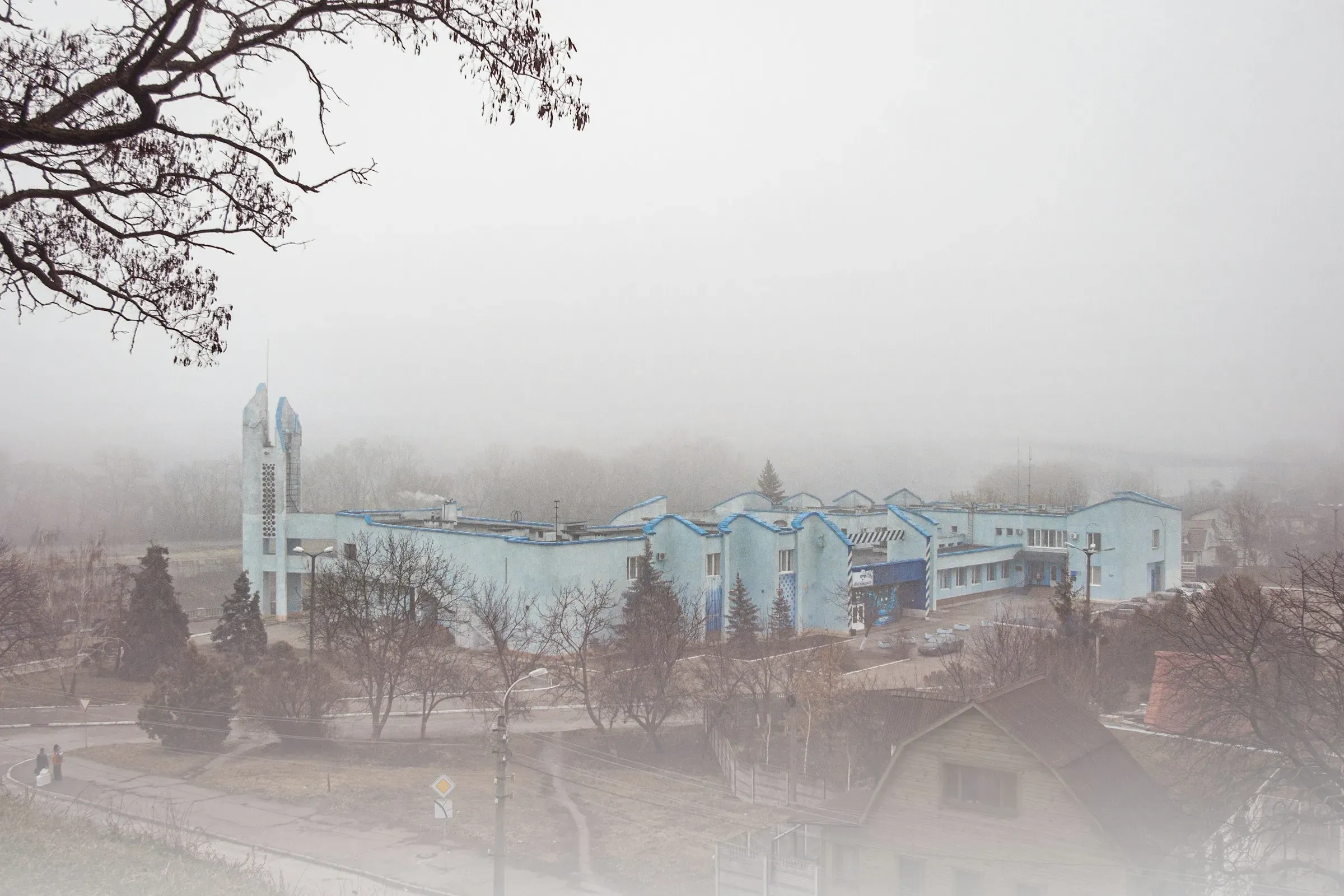 A low industrial building in heavy fog, bare trees in front of it.