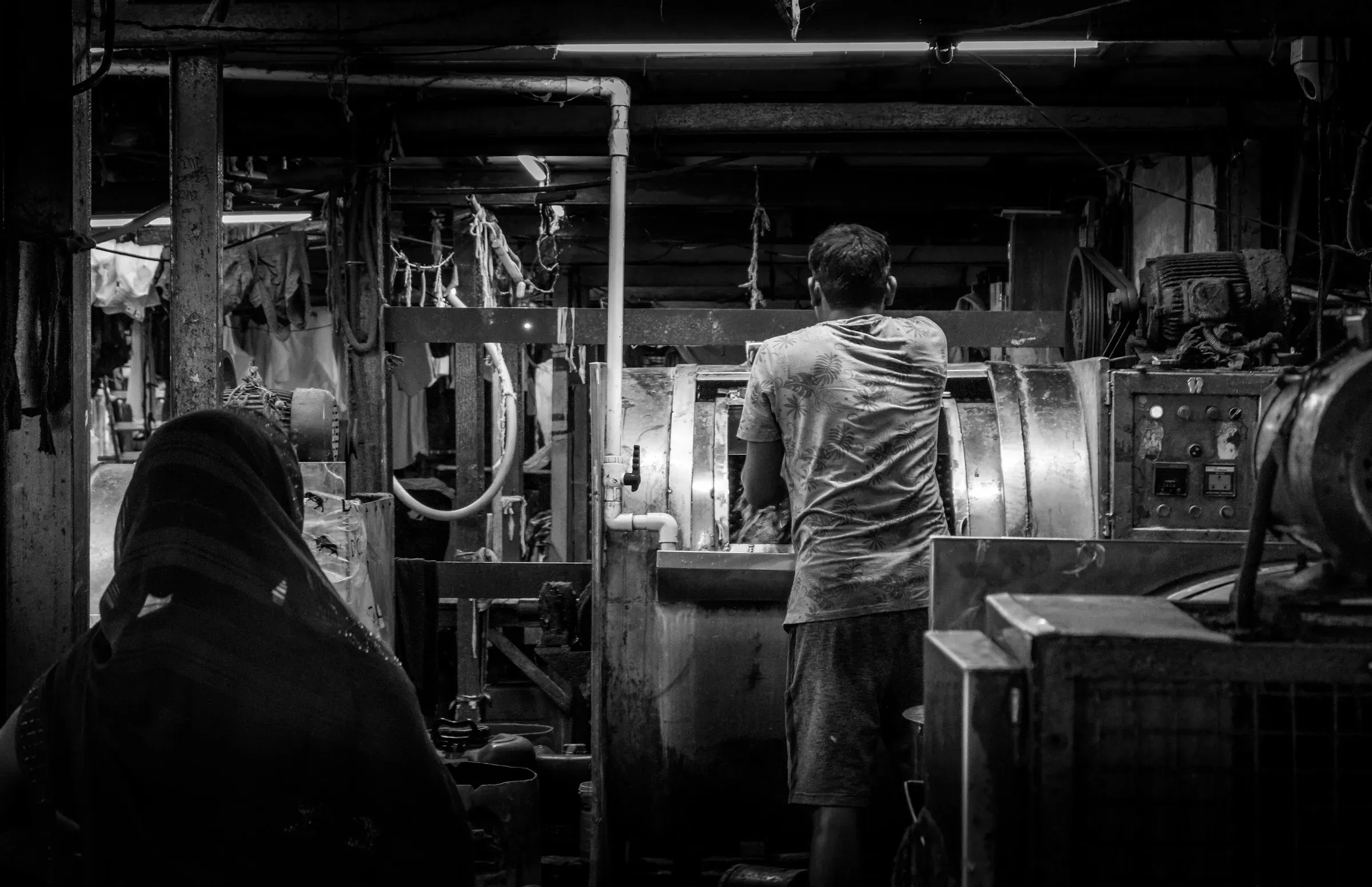 A dim workshop floor, one man at a machine and a second figure watching from the near corner.